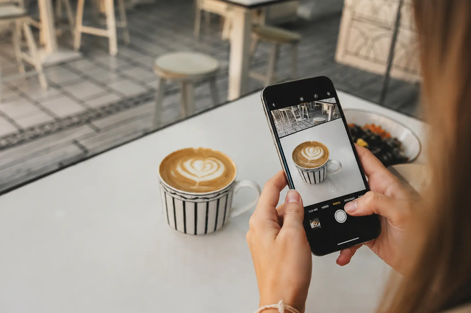 Person photographing a striped mug of latte art with a smartphone in a bright cafe