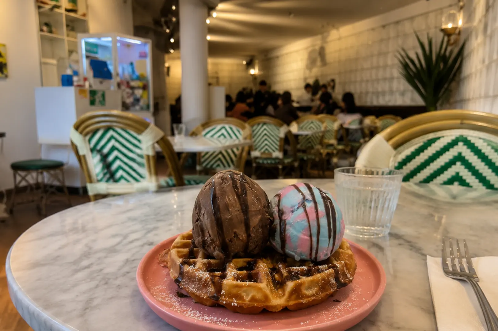 Waffle topped with chocolate and pastel ice cream scoops on a pink plate at a trendy cafe with green chevron chairs