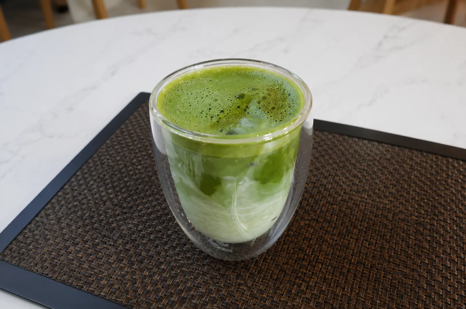 Iced matcha latte in a double-walled glass cup on a woven placemat at a marble table