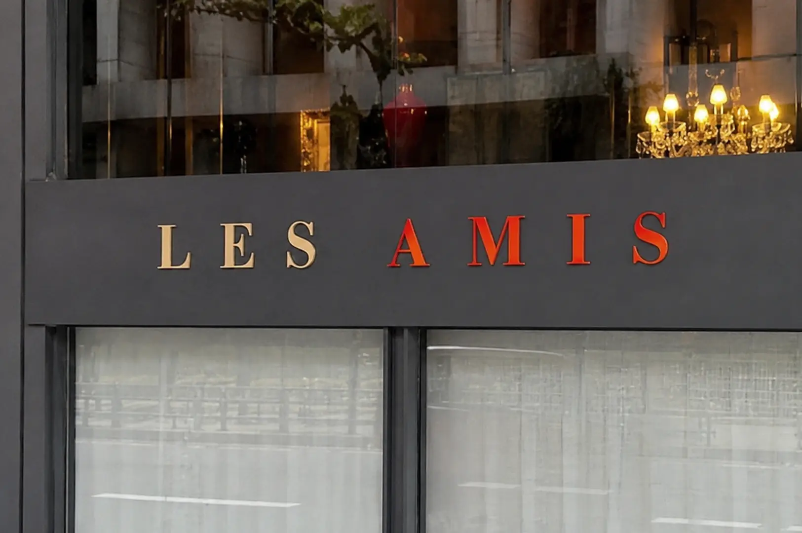 Close-up of the Les Amis restaurant sign featuring gold and red lettering on a dark facade with a chandelier visible inside