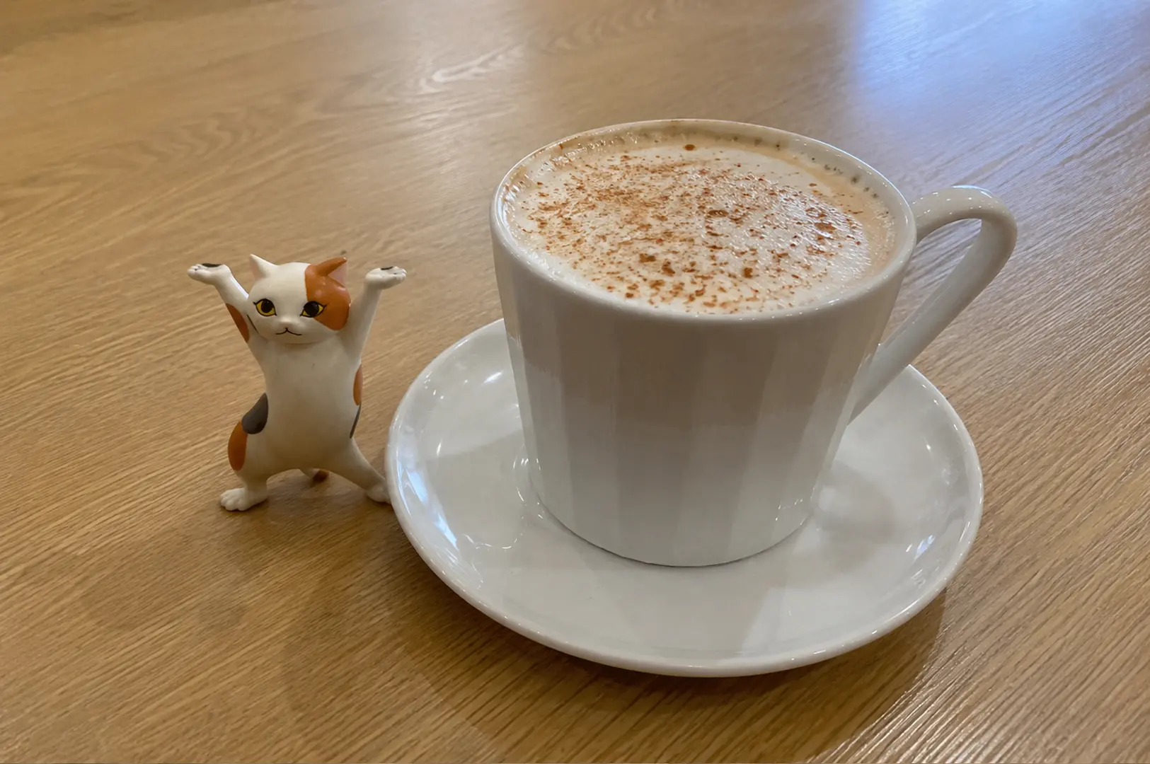 A frothy cappuccino topped with cinnamon sits on a wooden table next to a small figurine of a cheering calico cat