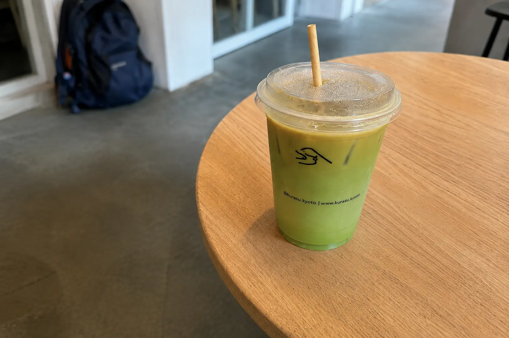 An iced dirty matcha drink in a Kurasu branded cup with the cafe's logo and website sits on a round wooden table