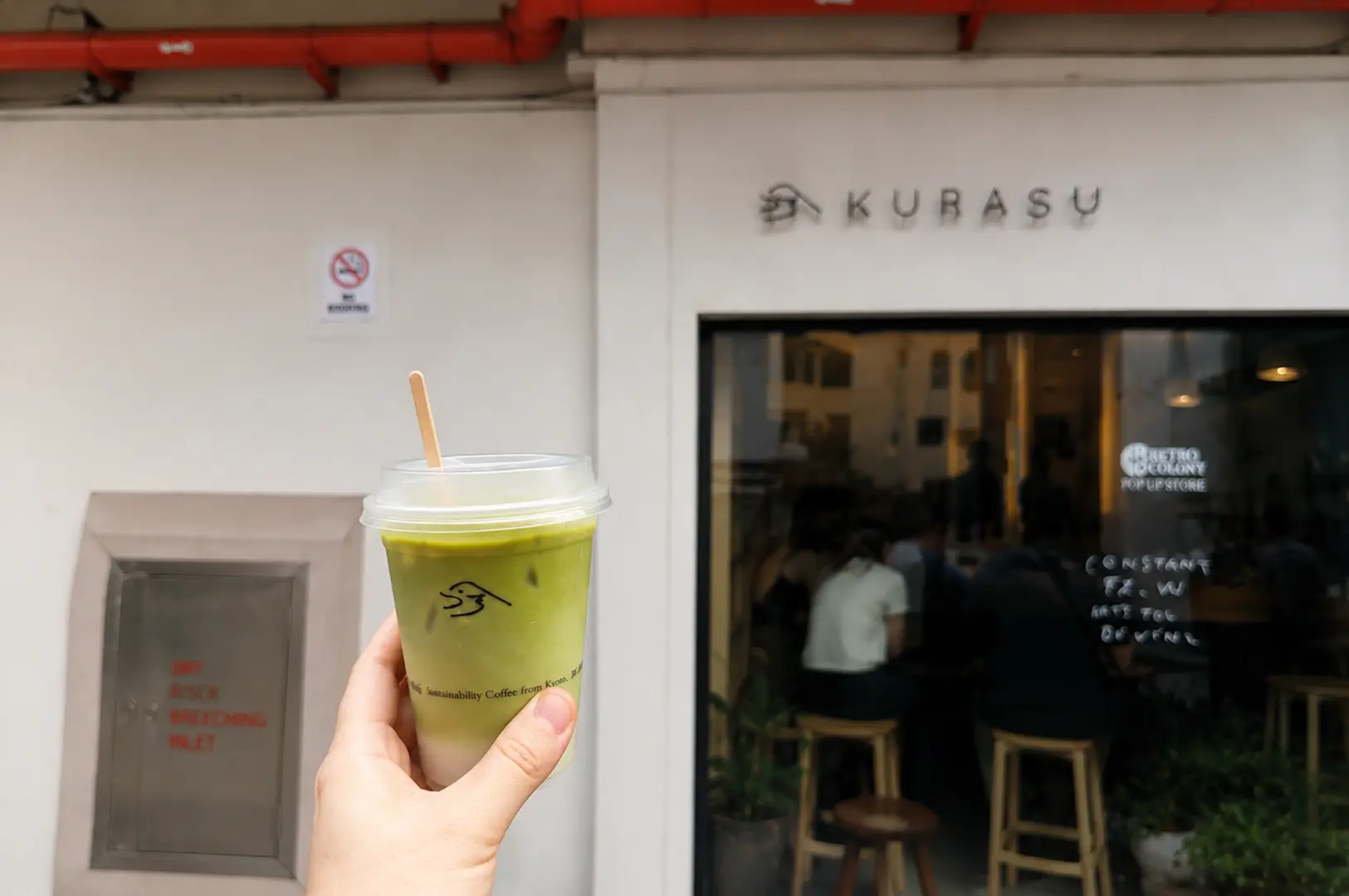 Hand holding an iced matcha drink in front of the Kurasu cafe storefront