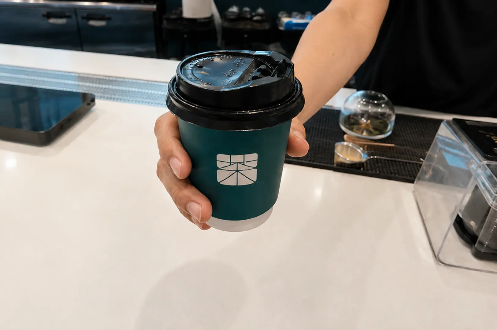 Staff serving a a teal Matchaya paper cup with a black lid over a white cafe counter