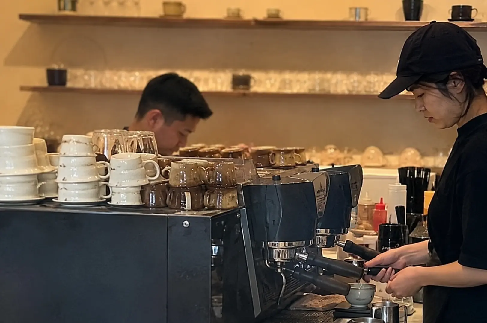 Baristas prepare drinks behind an espresso machine lined with stacked ceramic cups at Kurasu