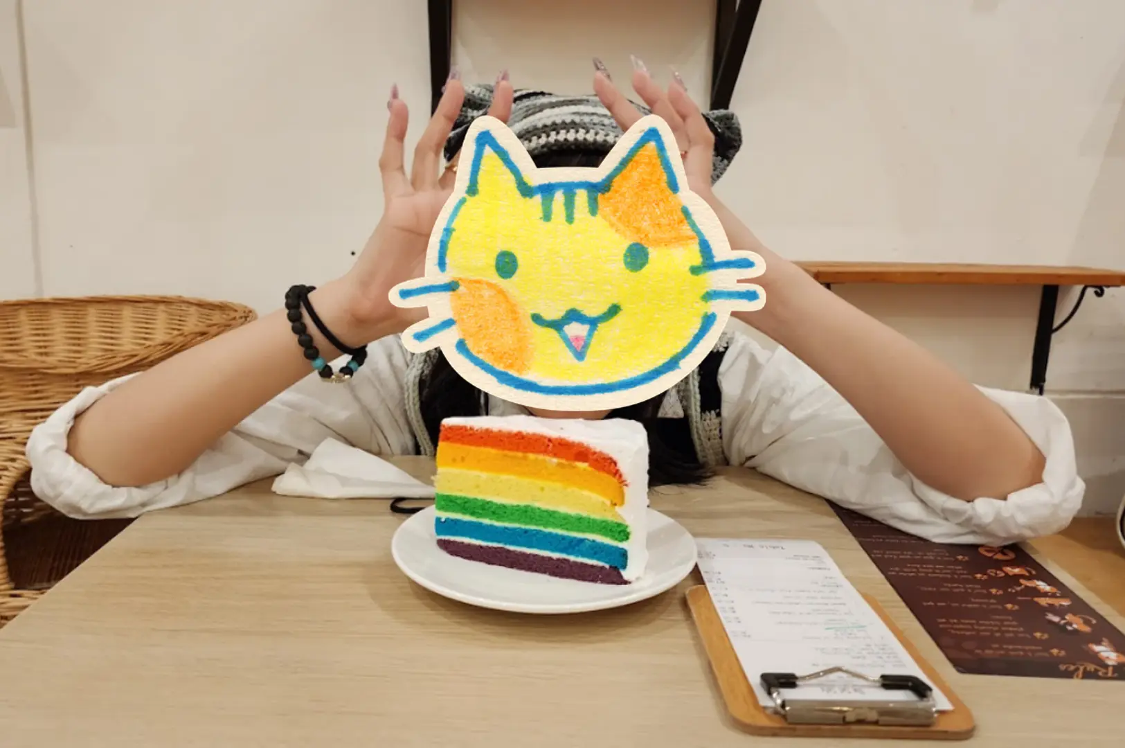 A visitor holds a colorful hand-drawn cat face cutout in front of a slice of rainbow layer cake