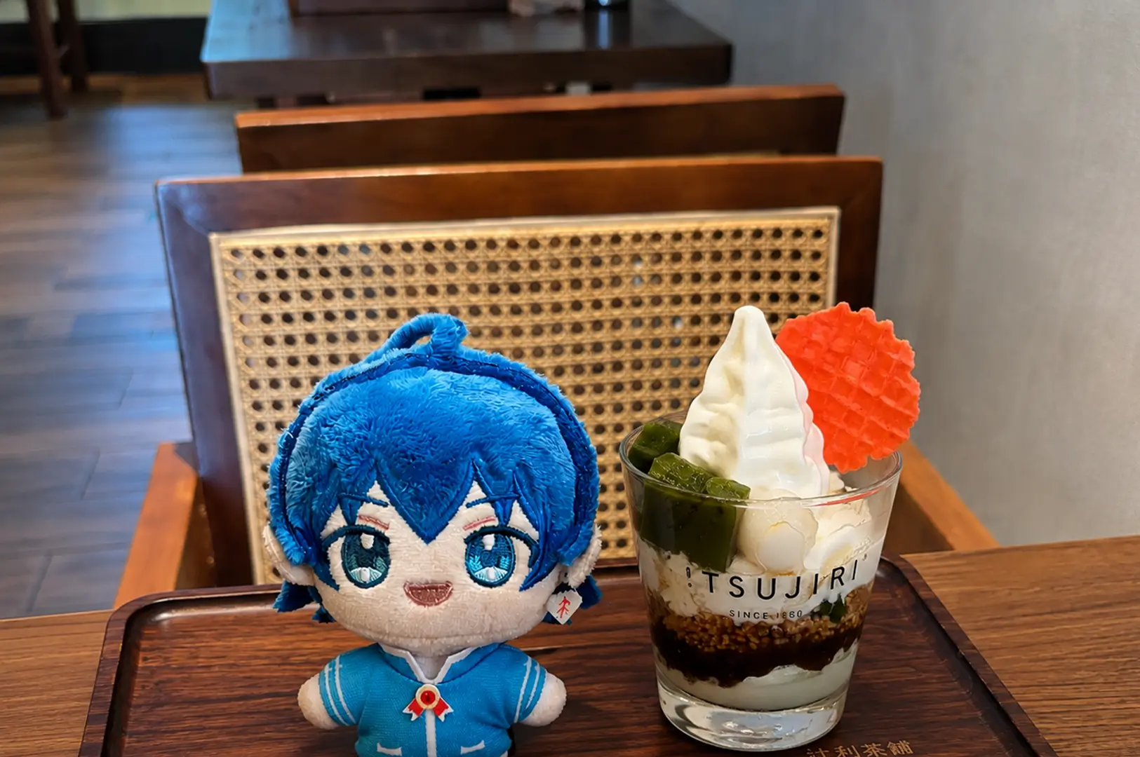 Blue anime plush doll sitting next to a Tsujiri matcha parfait with soft serve ice cream and a red wafer on a wooden tray