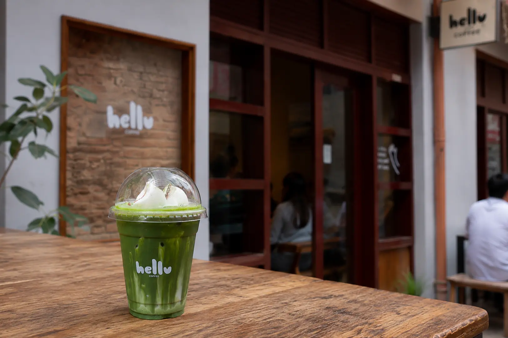 Iced matcha latte with cream topping in a plastic cup on a wooden table outside Hellu Coffee storefront