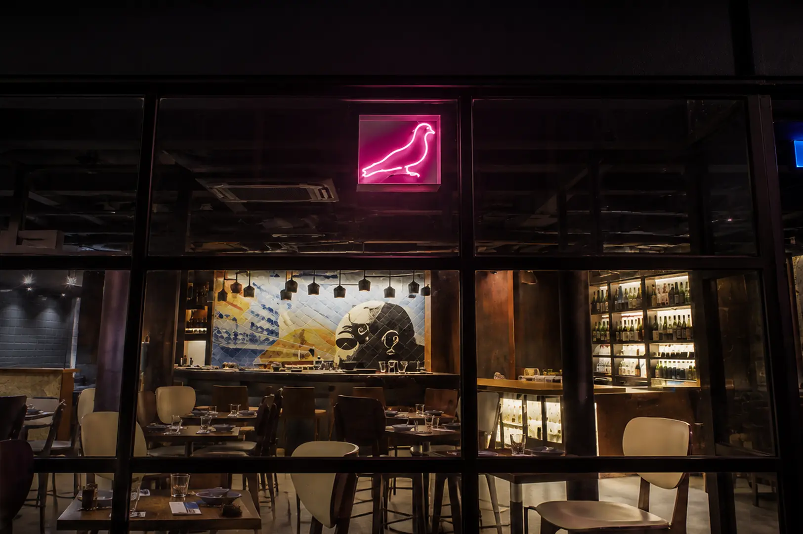 Nighttime view of a stylish izakaya through glass windows, featuring a bright pink neon bird sign, artistic wall mural, and shelves of sake bottles