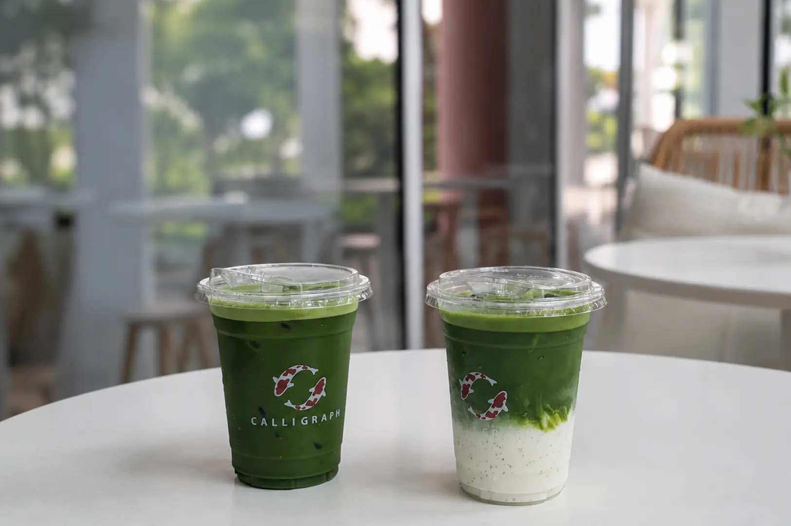 Two iced matcha drinks in green cups with Calligraph logo on a white table, one layered with a milky chia seed base