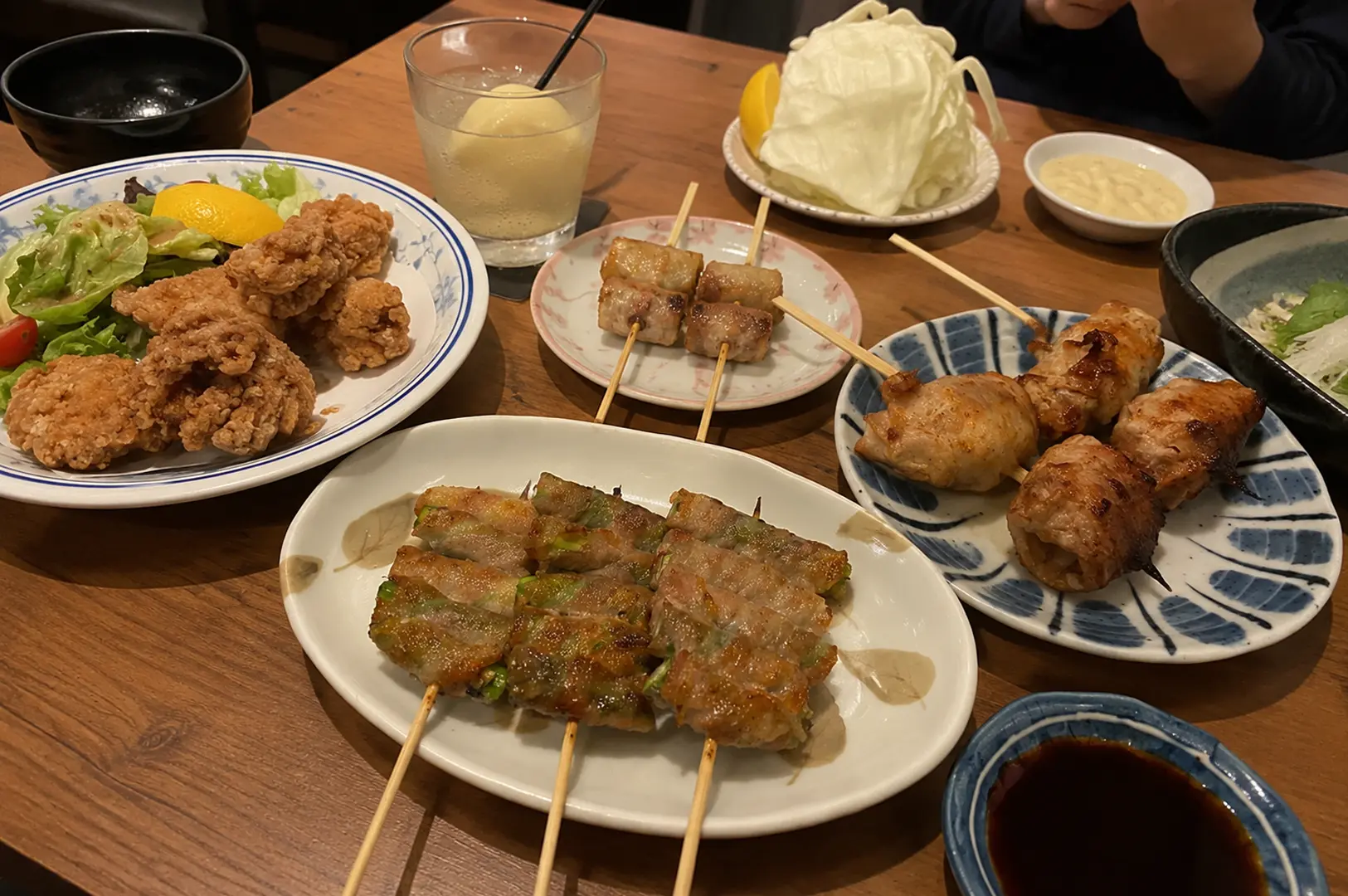 Wooden table spread featuring various izakaya dishes including yakitori skewers, bacon-wrapped rolls, fried chicken, and side dishes