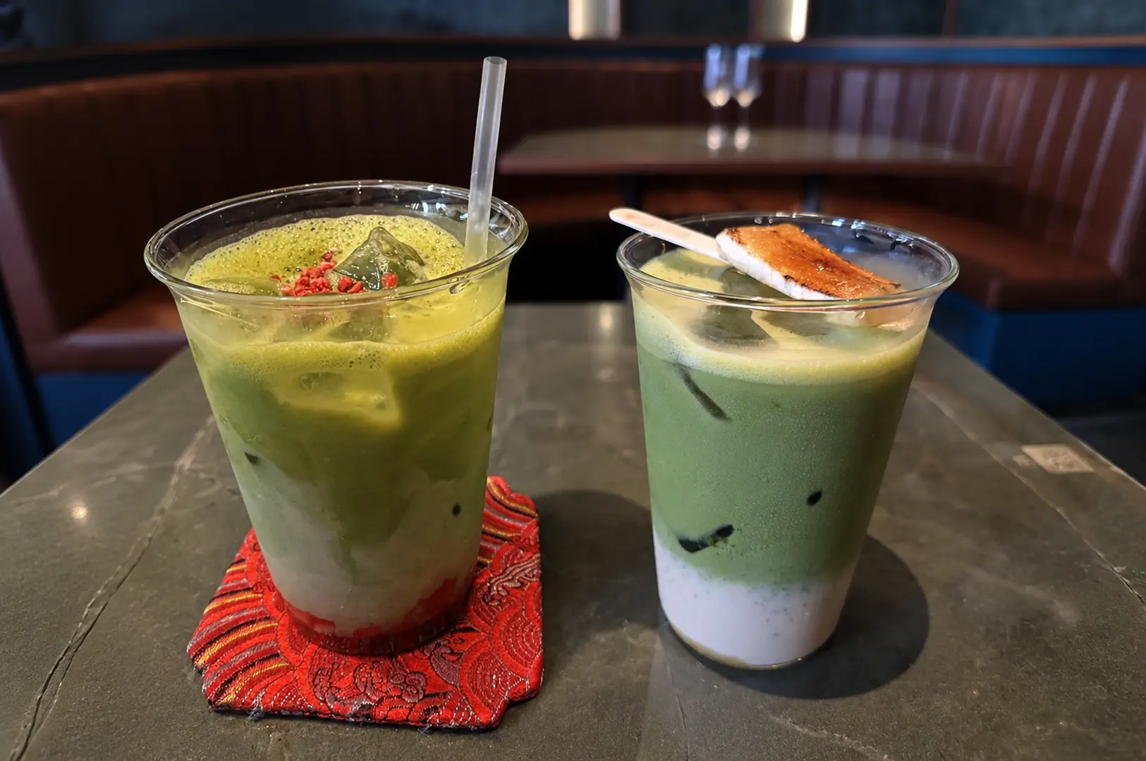 Cafe Yasuragi's two iced matcha drinks on a dark table, one with a red syrup base and sprinkles, the other with a marshmallow garnish