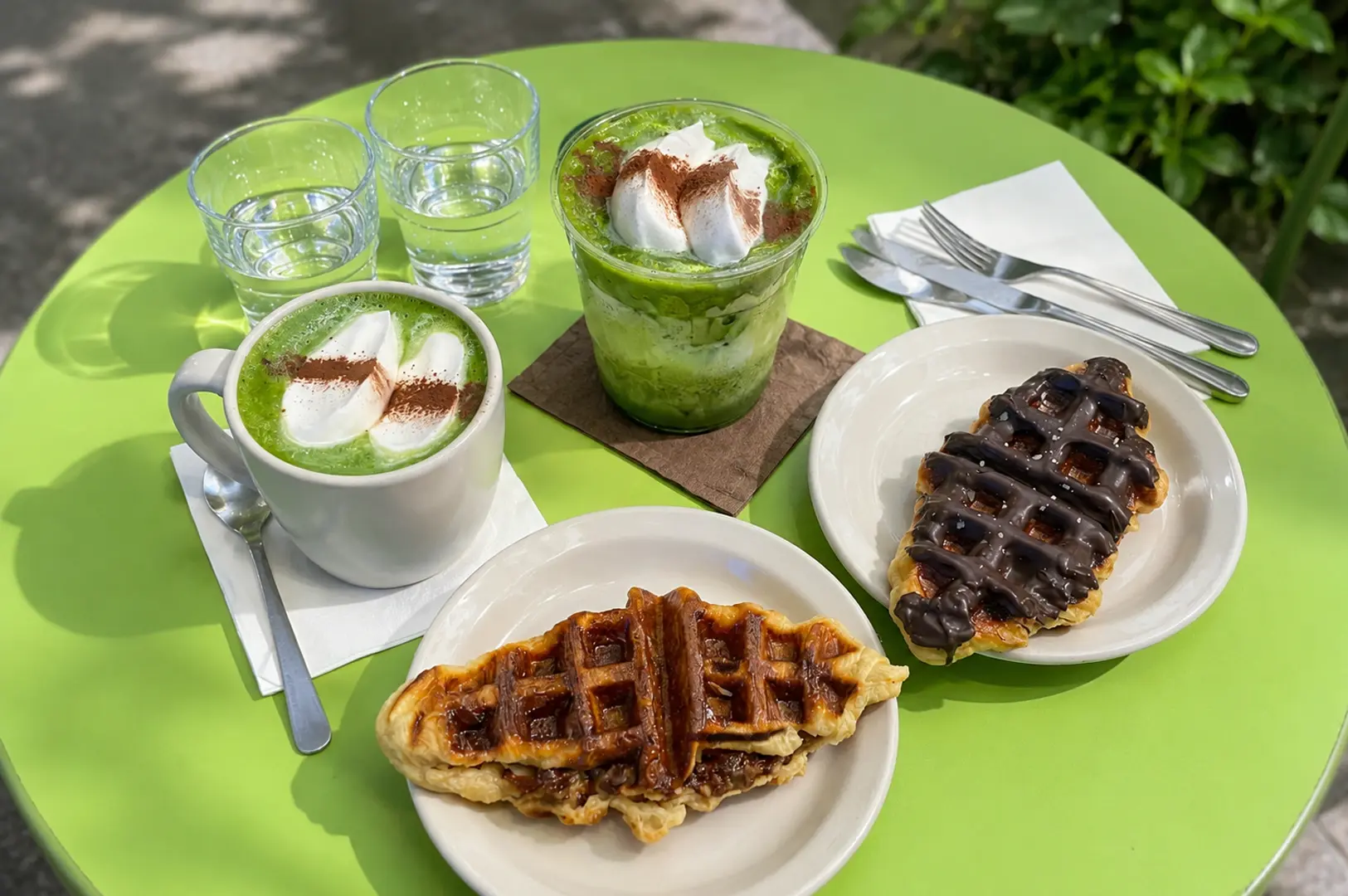 Bright green table set with hot and iced matcha lattes topped with cream and two waffles, one drizzled with chocolate