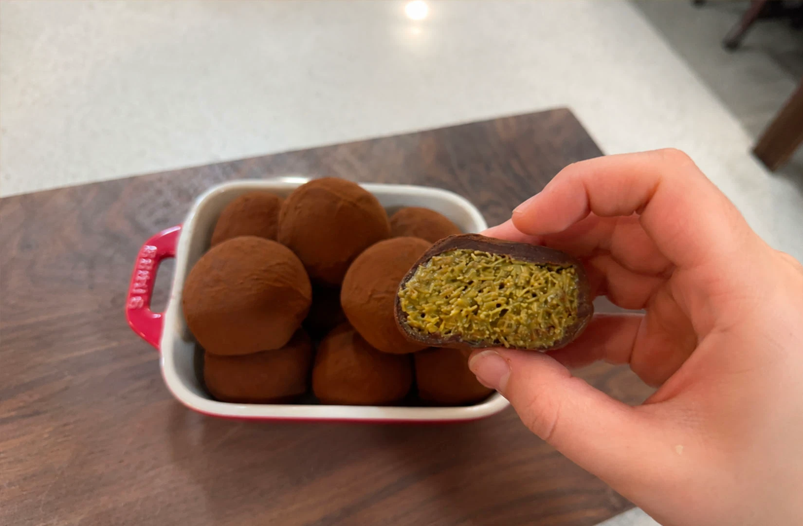 A hand holds a chocolate truffle cut in half to reveal a green pistachio and knafeh filling, with more truffles in a red dish behind it