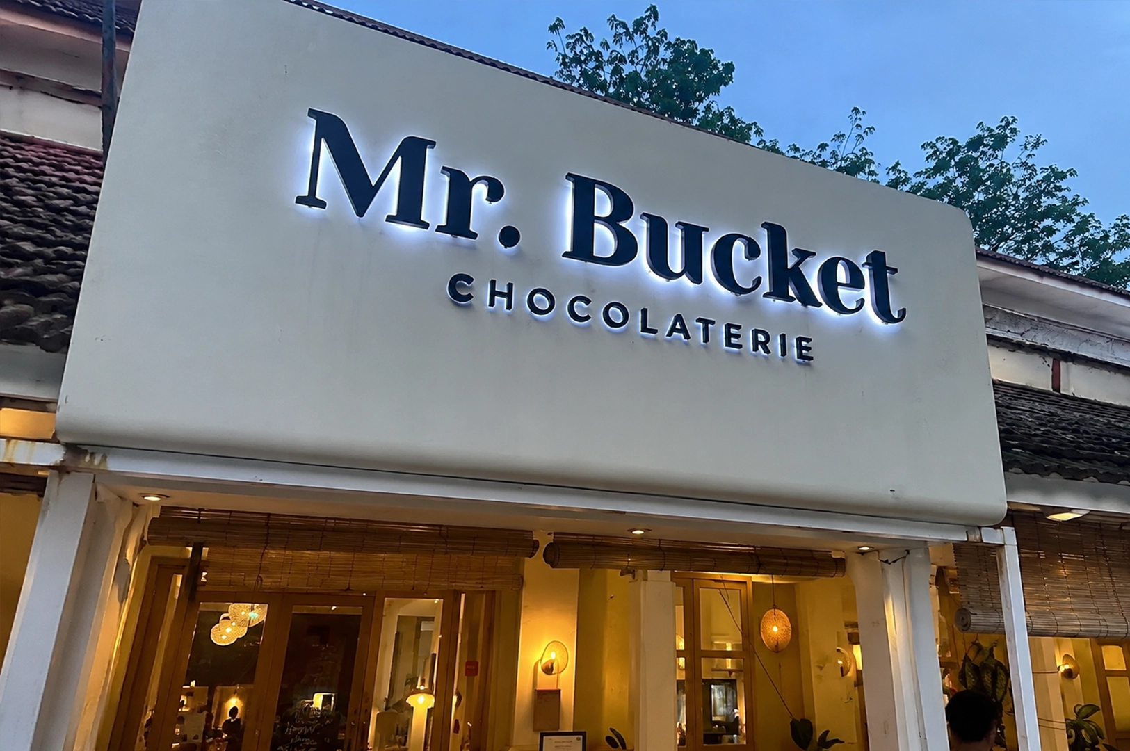 Evening view of Mr. Bucket Chocolaterie with its illuminated storefront sign welcoming visitors into the cozy chocolate café.