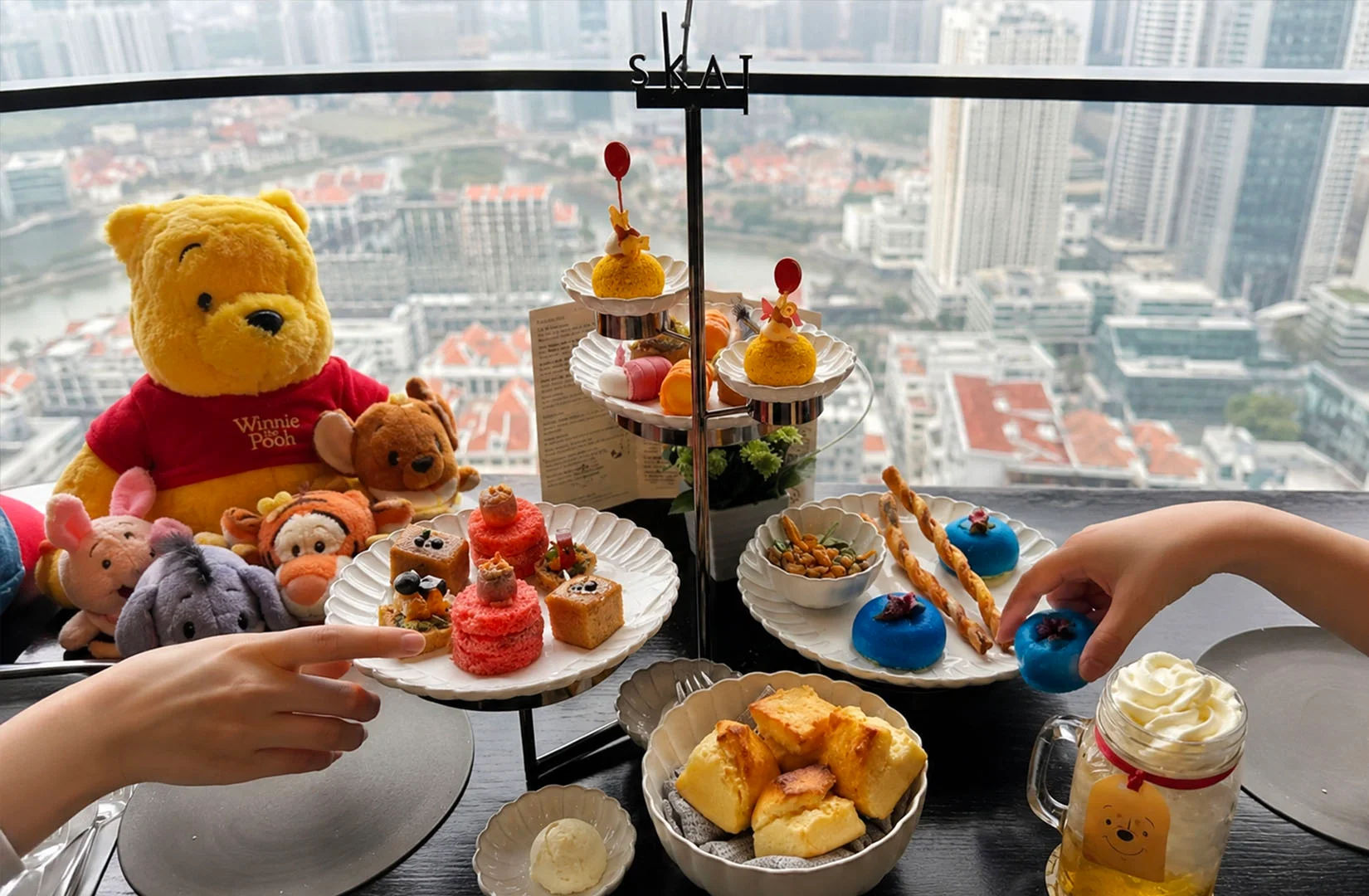 Winnie the Pooh plush sitting beside an elevated afternoon tea set with colorful pastries overlooking a city skyline through a high-rise window.