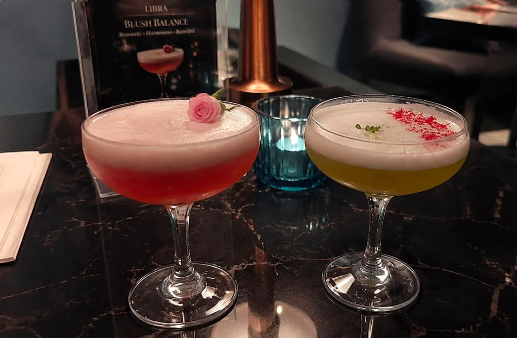 Two astrology-inspired cocktails with foamy tops and floral garnishes.