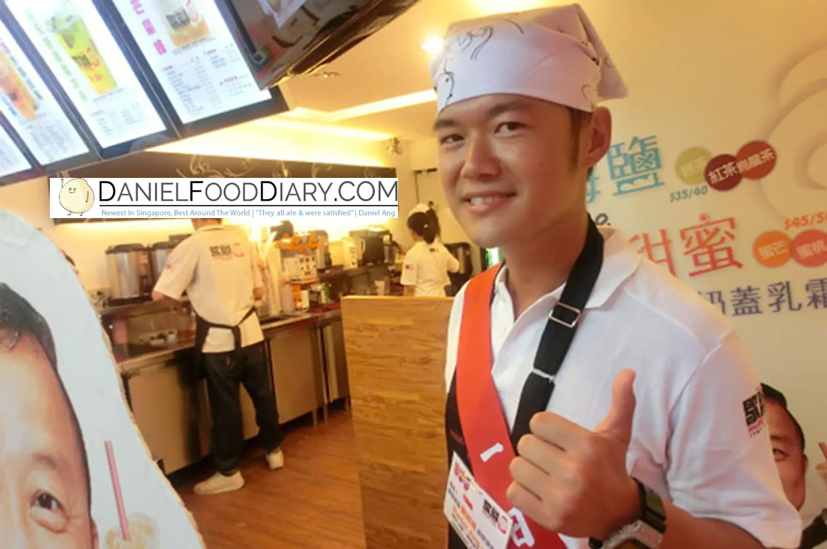 Daniel Ang giving a cheerful thumbs up while wearing an apron and bandana inside a bustling bubble tea shop