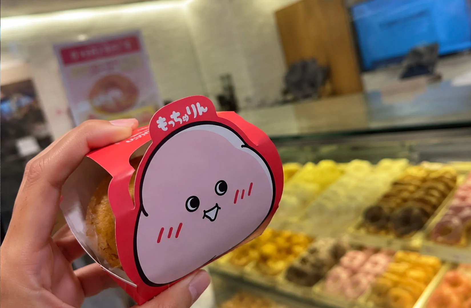 Hand holding a Mister Donut Mocchurin donut in its cute packaging with a blurred display of fresh donuts in the background.