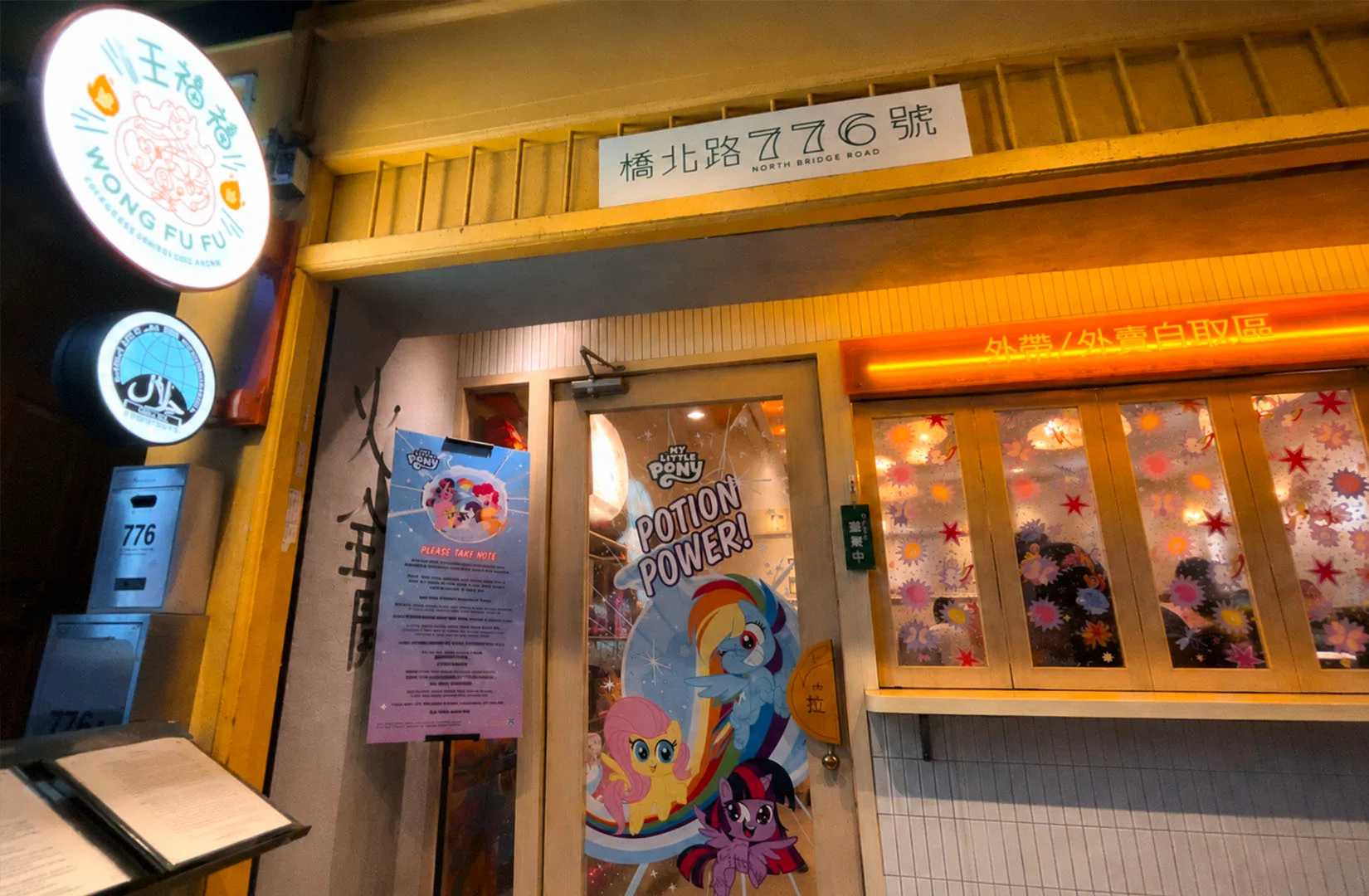 Bright storefront entrance with My Little Pony themed door graphics and signage, featuring colorful character posters and warm exterior lighting.