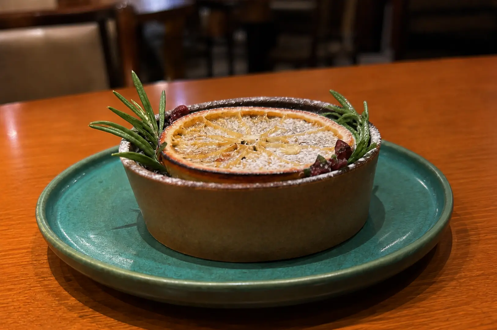Close-up of a handcrafted chocolate passionfruit tart topped with a dried citrus slice, rosemary sprigs, and a light dusting of powdered sugar on a green ceramic plate.