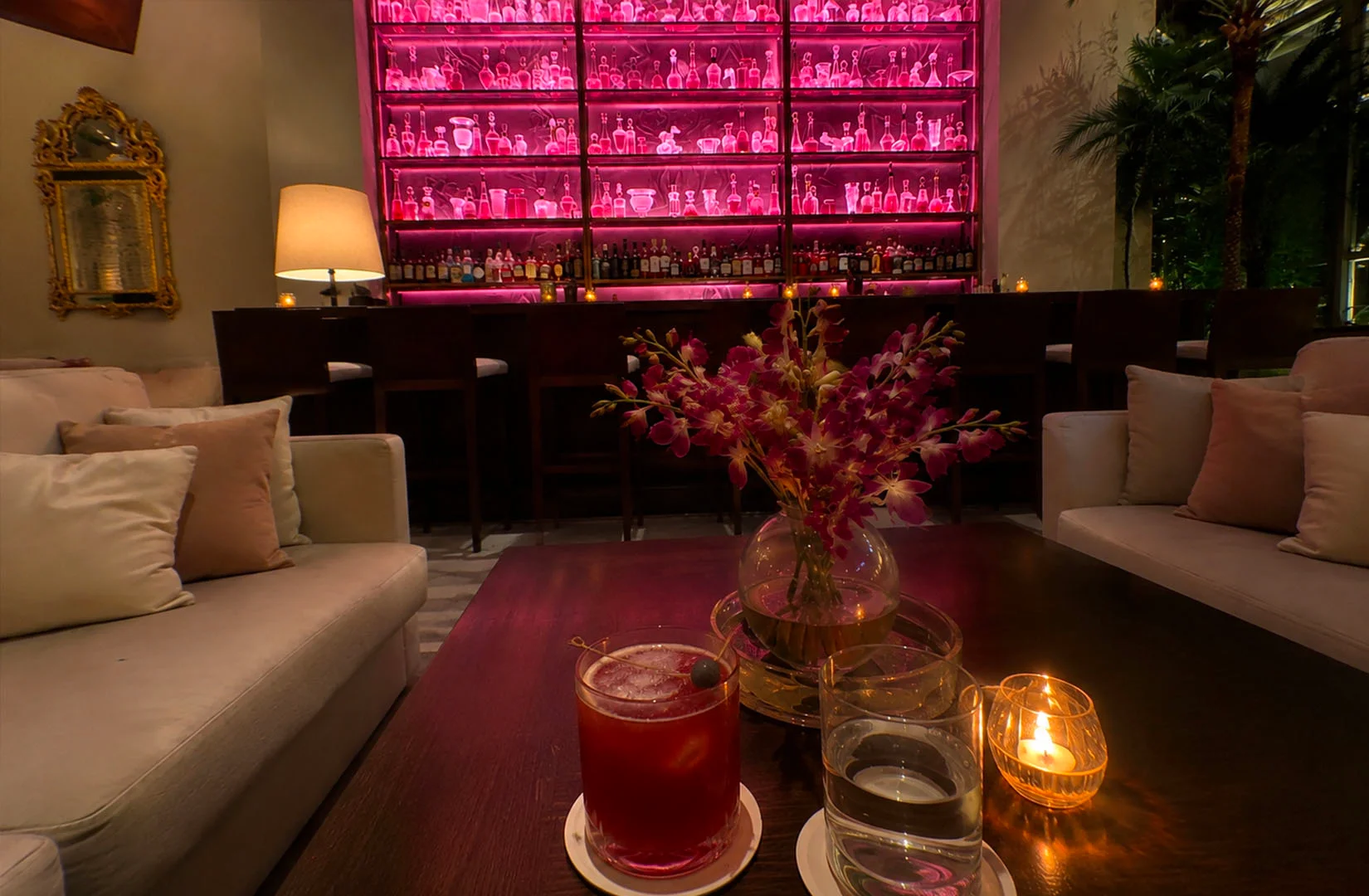 Cozy upscale lounge bar with pink backlit shelves of bottles, candle-lit table, and cocktails in a dim romantic interior setting.
