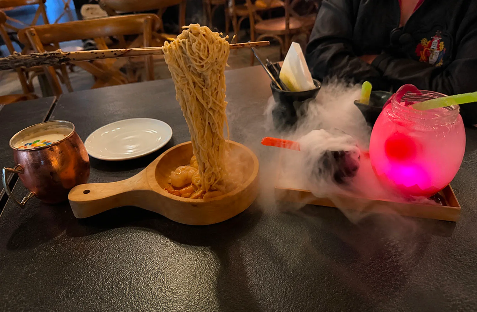 Ramen noodles lifted with chopsticks beside a smoky glowing drink in a restaurant setting.