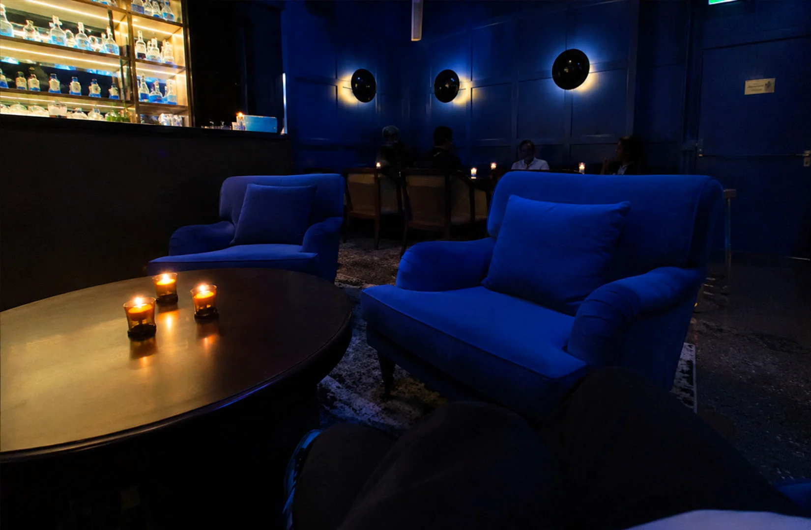 Dark blue intimate cocktail lounge with plush armchairs, candle-lit tables, and a back bar display creating a moody speakeasy atmosphere.