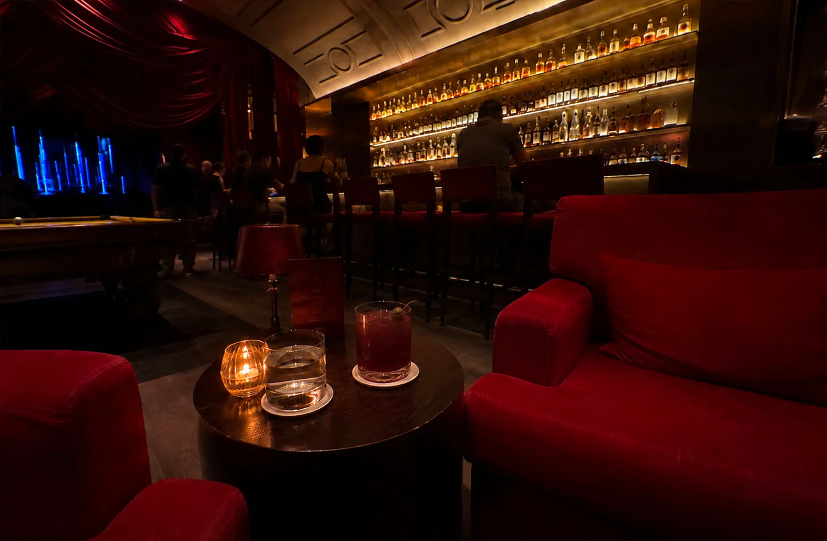Dim luxury lounge bar with red velvet seating, glowing backlit shelves of bottles, and cocktails on a table in a warm moody atmosphere.
