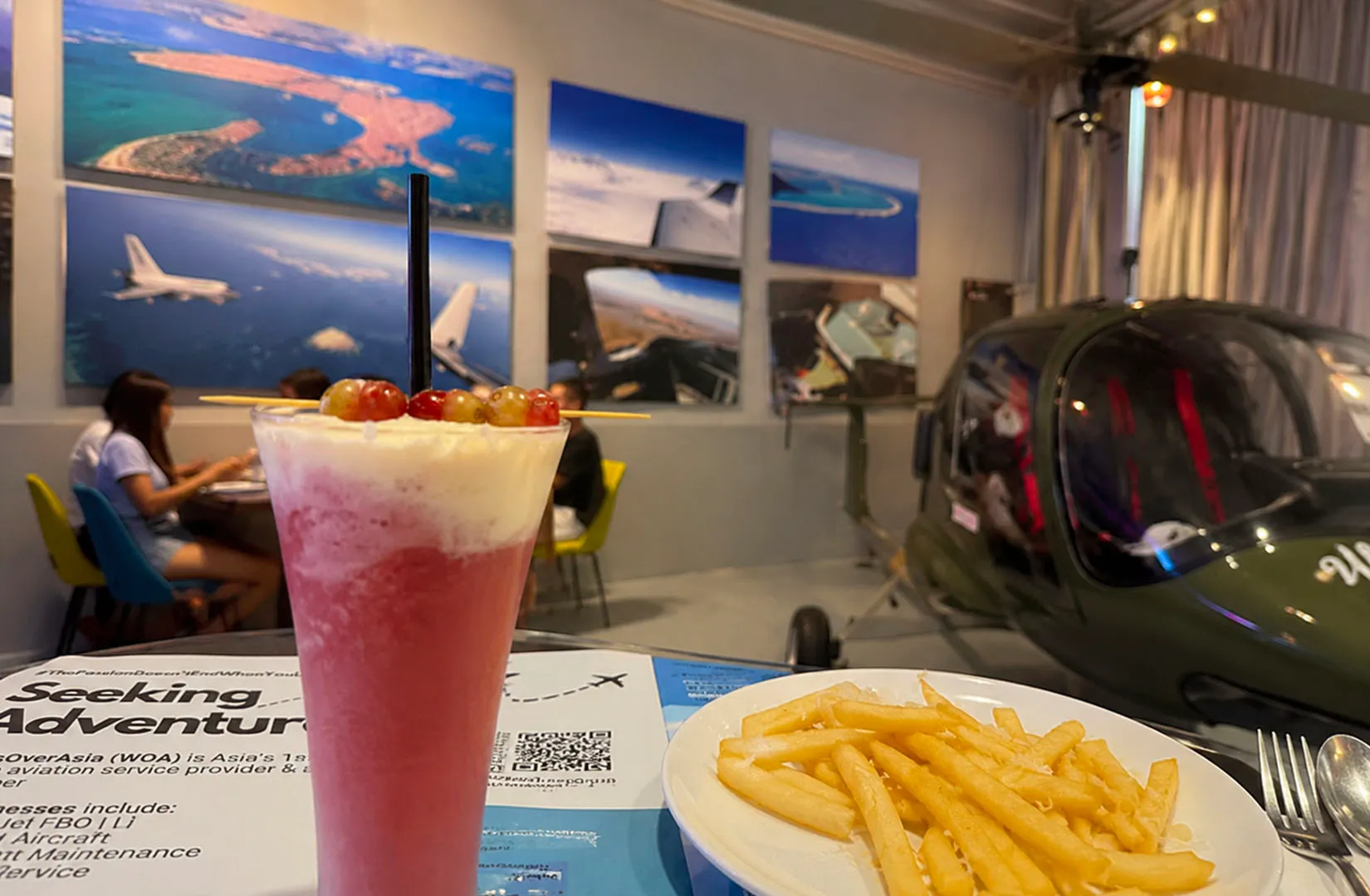 Airplane-themed cafe with pink drink topped with foam and grapes, and fries on the table with aviation decor in the background.