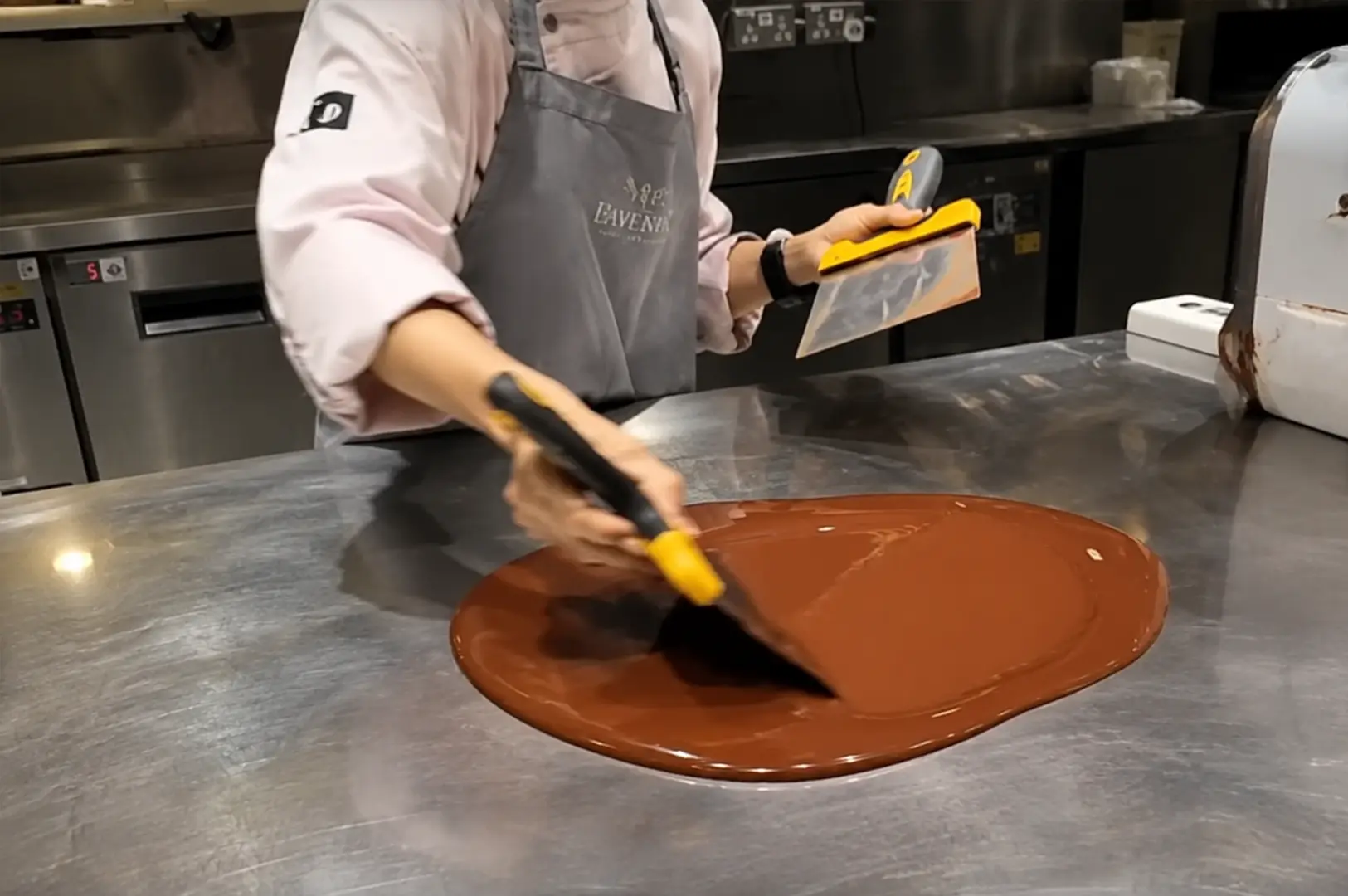 A chocolatier carefully tempers and spreads melted chocolate across a stainless steel worktable during the bean-to-bar chocolate-making process.