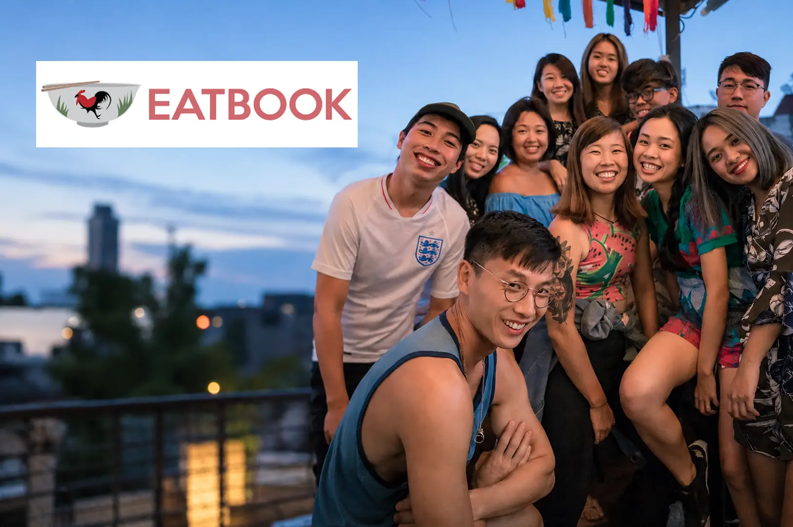 The cheerful Eatbook team gathered together on a rooftop terrace at dusk, with colorful decorations overhead and city buildings in the background