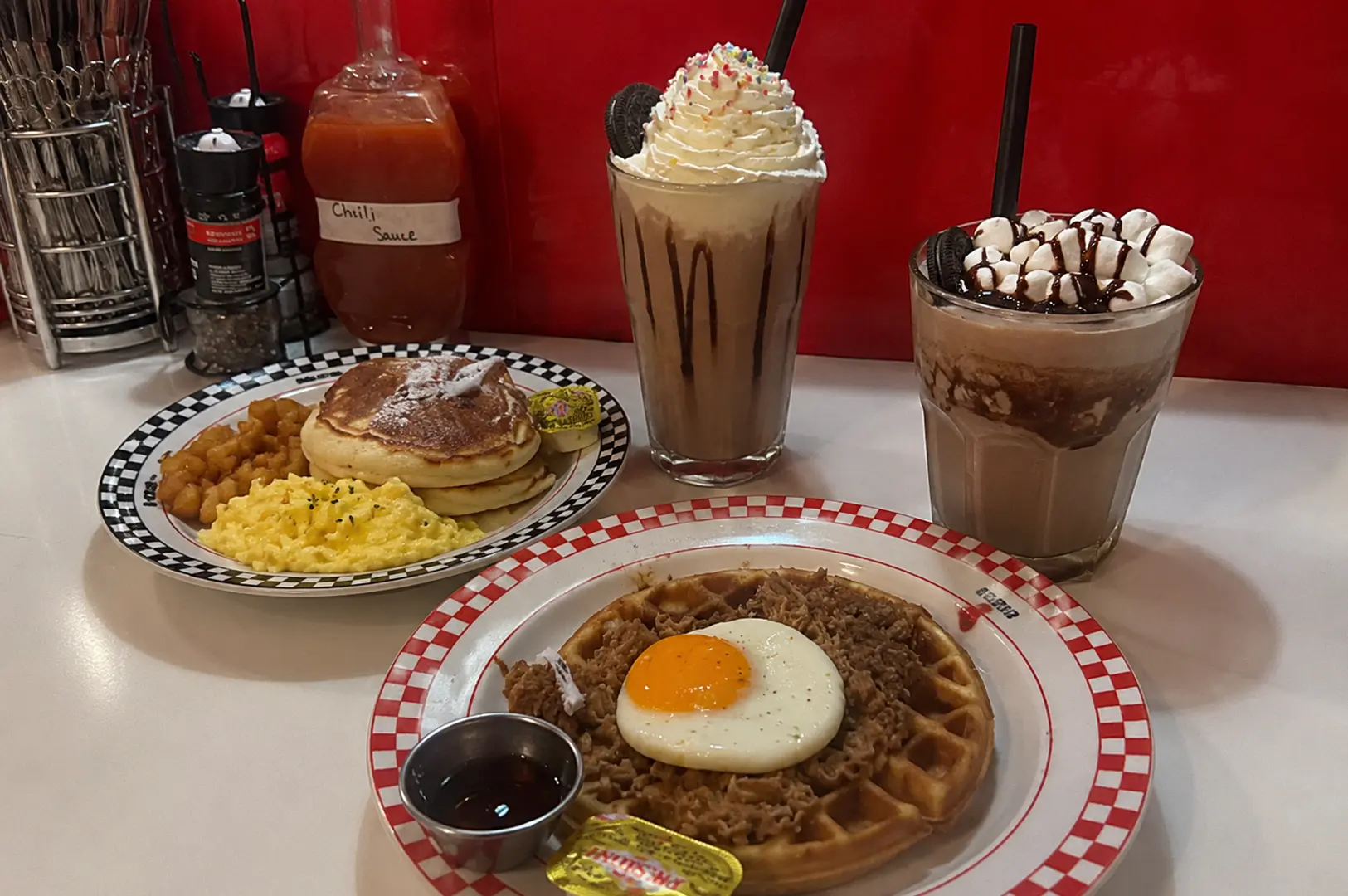 Breakfast spread at Joji’s Diner featuring pulled meat waffles with a sunny-side-up egg, pancakes, scrambled eggs, and two chocolate drinks.
