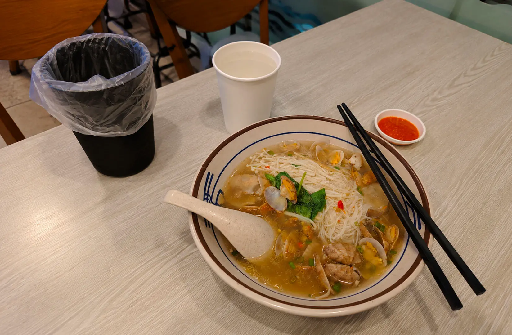 Clam bee hoon noodle soup with chili sauce and chopsticks in a casual eatery
