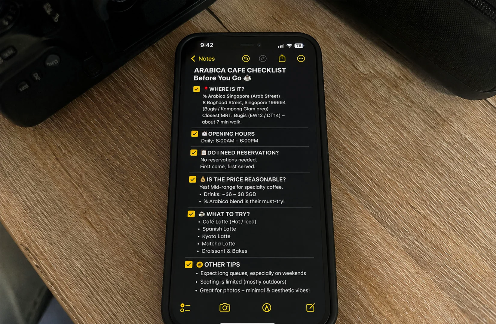 Smartphone showing a detailed “Arabica Cafe Checklist” note on a wooden desk with coffee shop planning info like location, hours, and drink recommendations