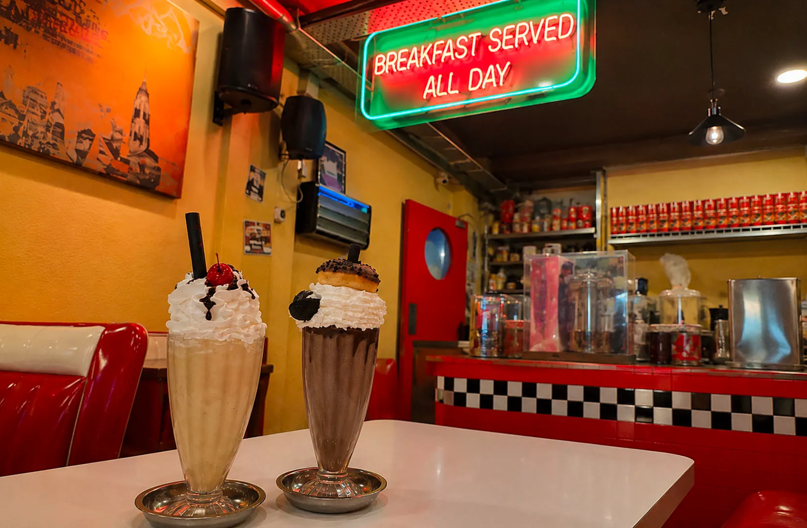 Milkshakes topped with whipped cream in a retro diner with neon “Breakfast Served All Day” sign.