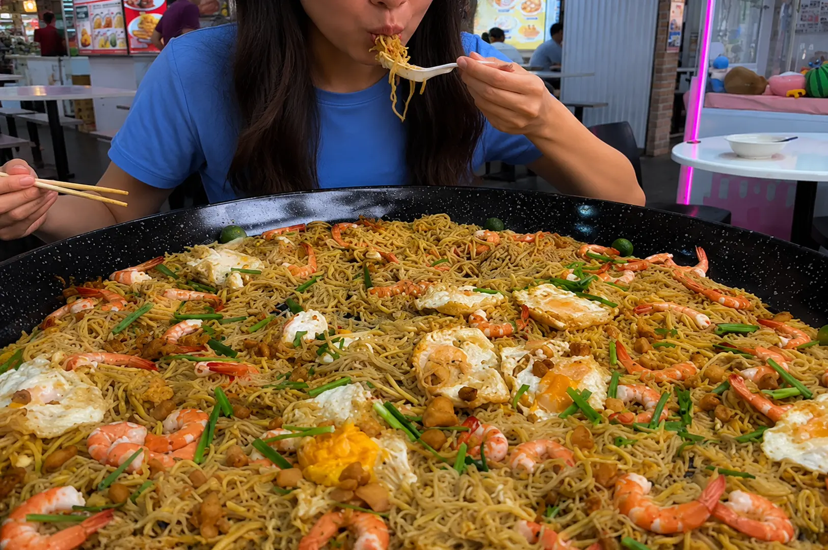 A diner in a blue shirt lifting noodles with a spoon and chopsticks from an enormous black wok filled with mee goreng topped with prawns, fried eggs, and spring onions at a hawker center