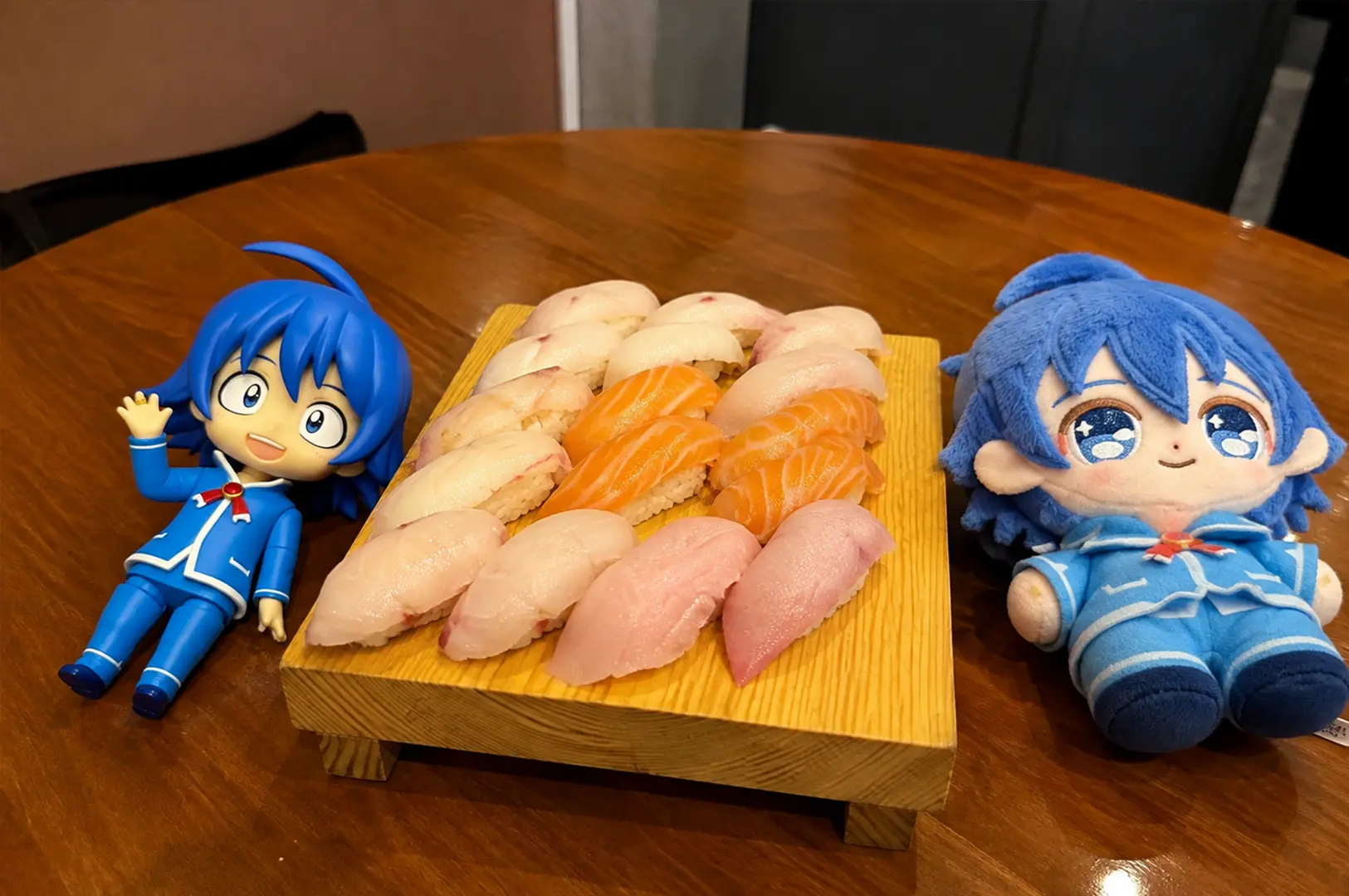 A wooden sushi platter filled with assorted nigiri, including salmon and pale white fish, displayed between two blue-haired anime figures on a dining table.