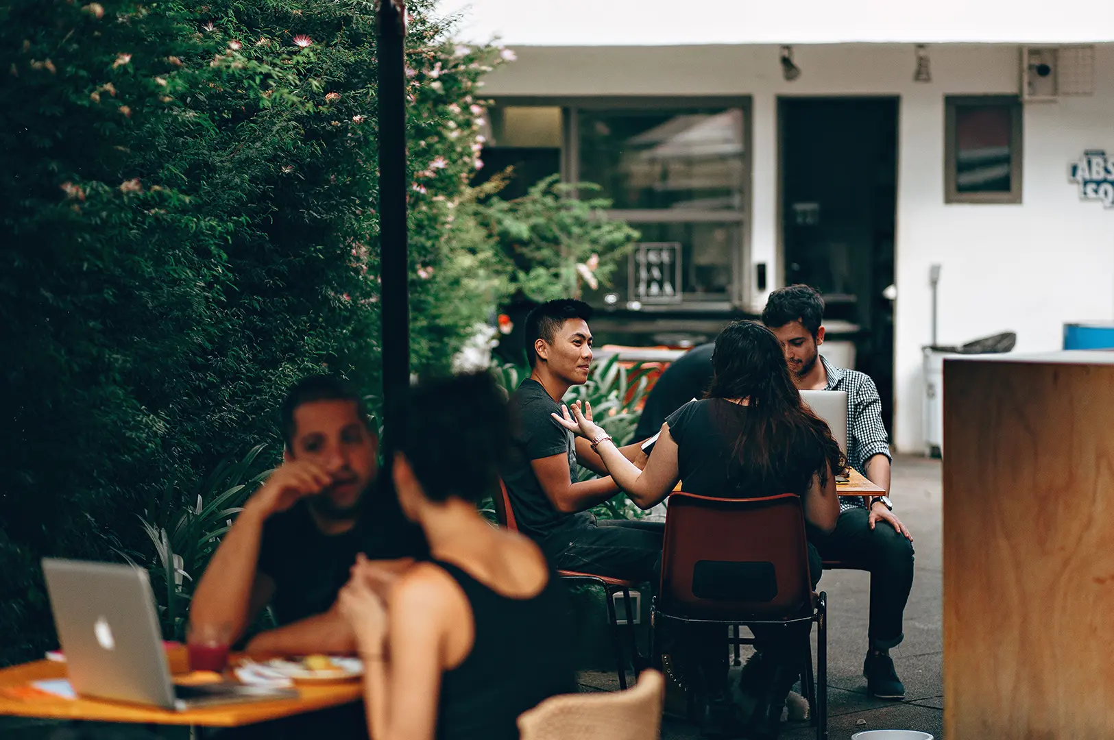 People working and chatting with laptops in a busy outdoor cafe during the day.