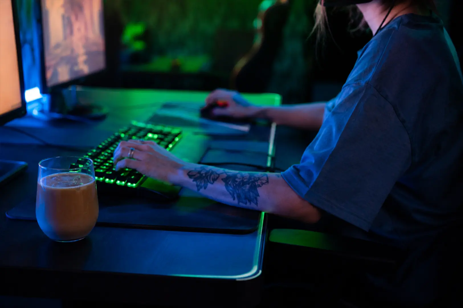 A gamer using a desktop computer at a dark, neon-lit esports café, with multiple monitors, a gaming keyboard, and a drink on the desk.
