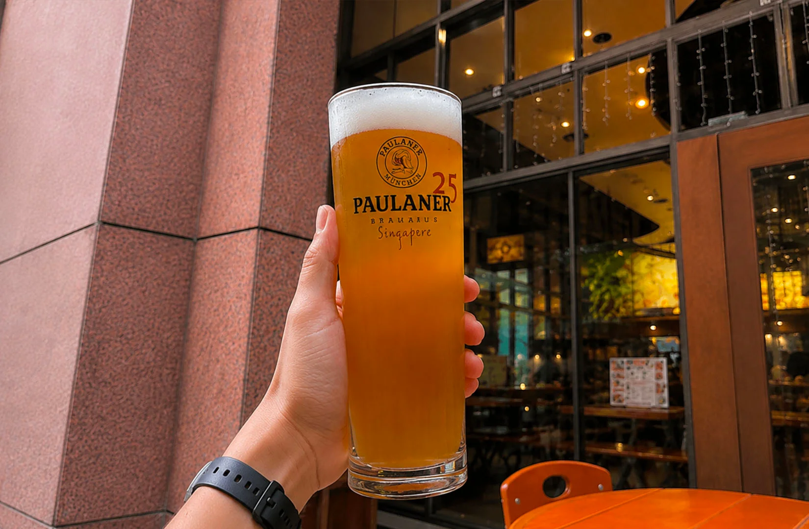 Freshly poured Paulaner wheat beer in a tall glass held outside Paulaner Brauhaus Singapore.