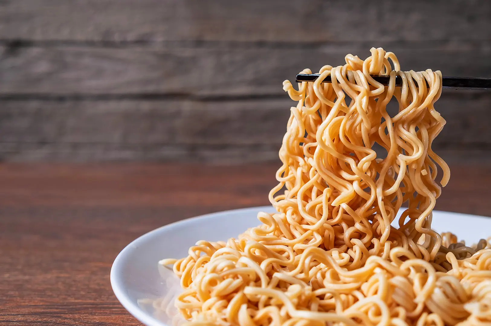 Plain cooked instant noodles lifted with chopsticks above a white plate.
