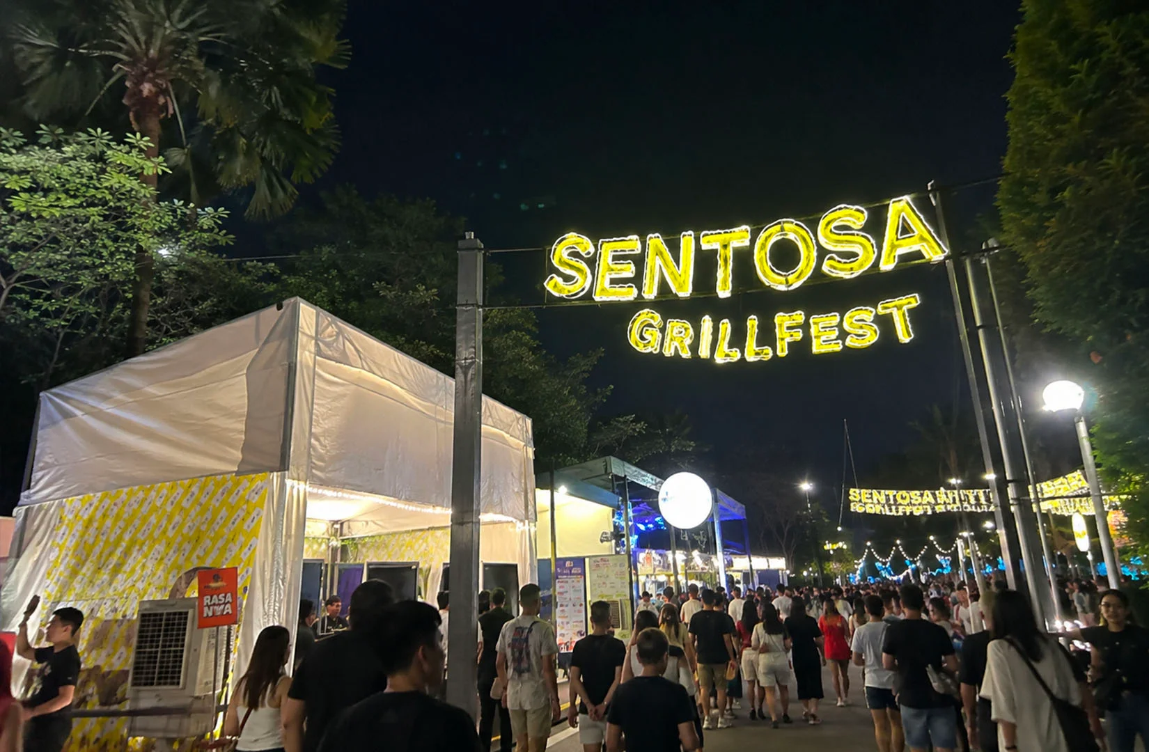Visitors walking through the brightly lit entrance of Sentosa GrillFest at night with food stalls on both sides.