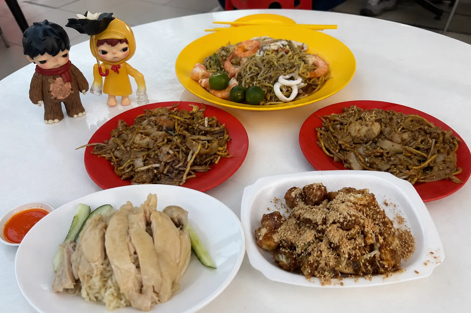 A spread of Singapore hawker classics, including seafood noodles, fried noodles, chicken rice and rojak, arranged with collectible figures.