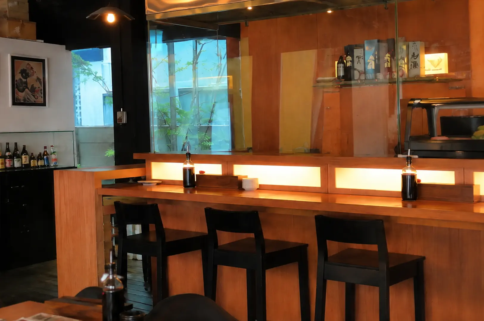 A warm, wood-paneled sushi bar with black chairs, individual dining counters, hanging pendant lights, and shelves displaying Japanese bottles at Standing Sushi Bar.