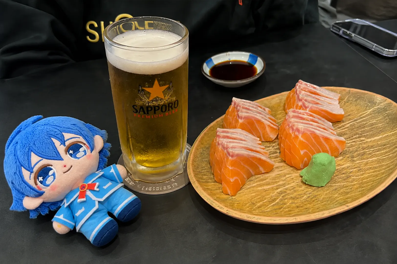 Thick slices of fresh salmon sashimi served on a wooden plate beside a glass of Sapporo beer, with a blue anime plush at Standing Sushi Bar.