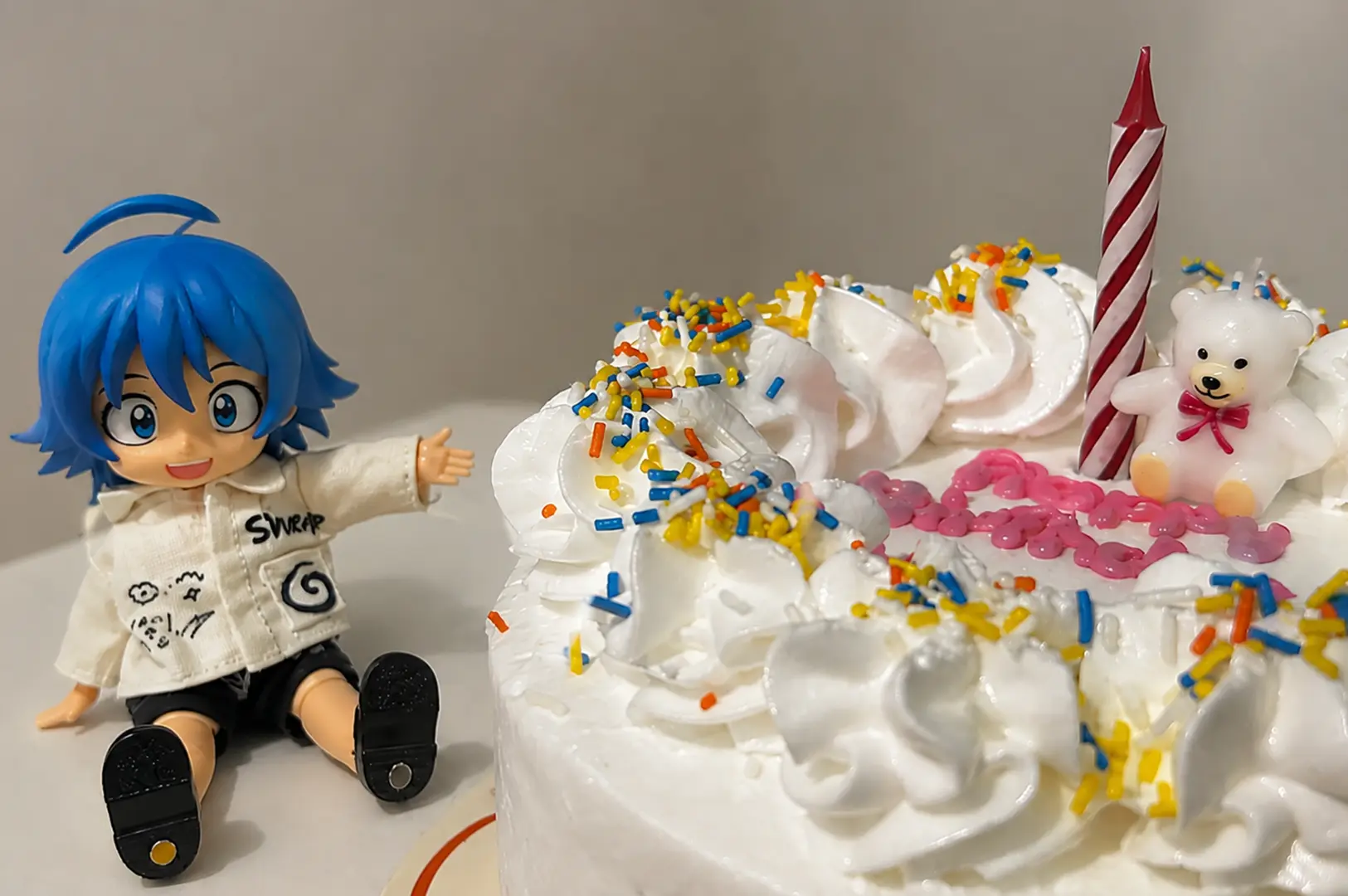 Blue-haired anime figure sitting beside a whipped cream birthday cake with sprinkles and a candle.