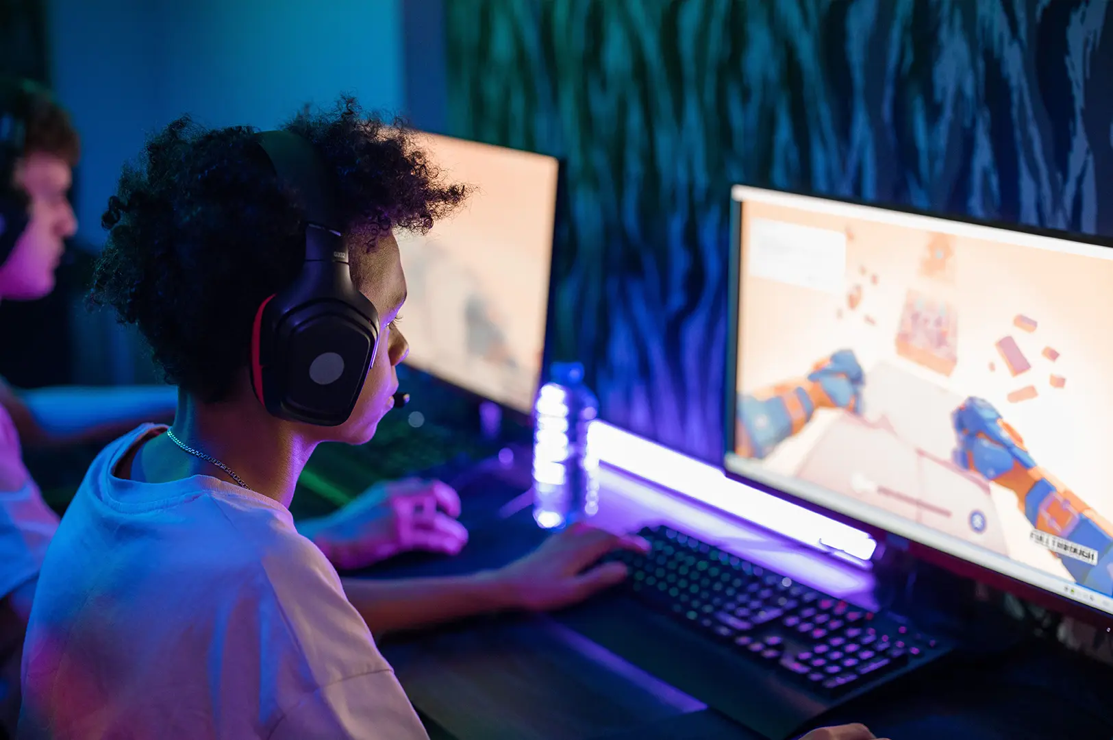 Two gamers playing on desktop computers in a colorful esports café, with one player wearing headphones and a game displayed on the monitor.