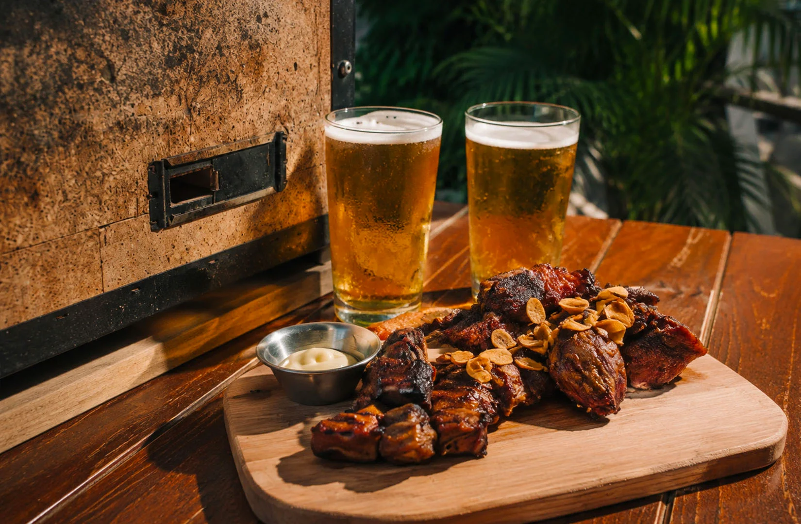Grilled barbecue meat topped with crispy garlic, served with two glasses of beer beside a charcoal oven.