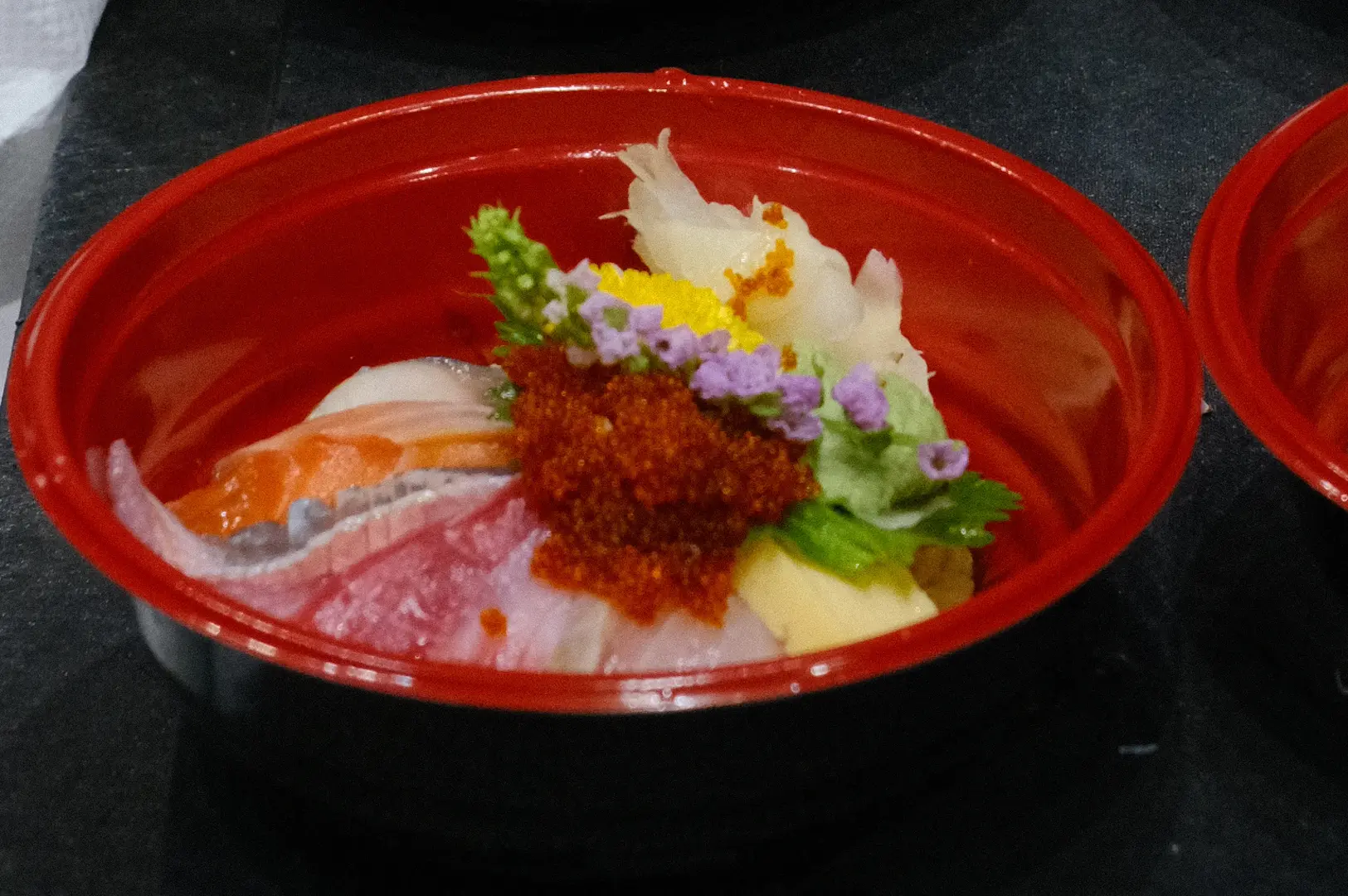 Red bowl of assorted sashimi topped with fish roe, garnishes, and edible flowers.